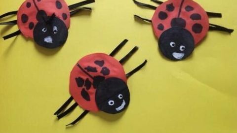 Drei bunte Marienkäfer aus Papier liegen auf einem gelben Hintergrund und lächeln fröhlich. Sie sind kreativ gestaltet.