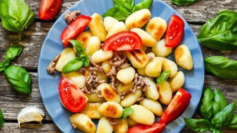 Eine bunte Gnocchi-Pfanne mit Hackfleisch und frischen Tomaten liegt auf einem blauen Teller, umgeben von Basilikumblättern.