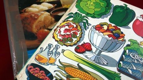 Ein Kochbuch mit bunten Illustrationen von Lebensmitteln liegt auf einem Tisch, umgeben von anderen Kochbüchern und Gerichten.