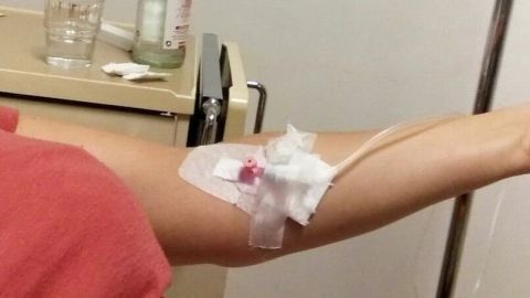 Ein Arm mit einer Infusion zur Thrombozytenspende liegt auf einer Liege, während im Hintergrund Wasser und Gläser stehen.
