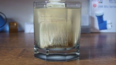 Ein Pinsel wird in ein Glas mit Lösungsmittel getaucht, um eingetrocknete Acrylfarbe zu entfernen. Der Tisch ist aus Holz.