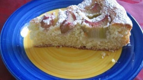 Ein Stück Rhabarber-Eierlikörkuchen liegt auf einem gelben Teller, umgeben von einem blauen Rand, auf einem Tisch.