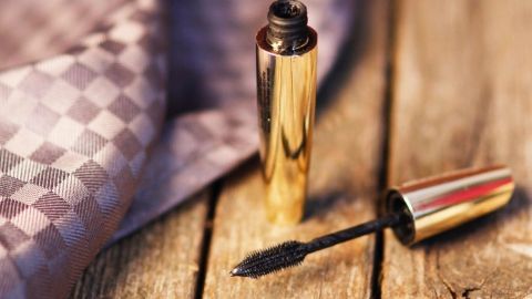 Ein goldener Mascara-Stift liegt offen auf einer Holzoberfläche, während der Pinsel auf einem eleganten Stoff ausgebreitet ist.