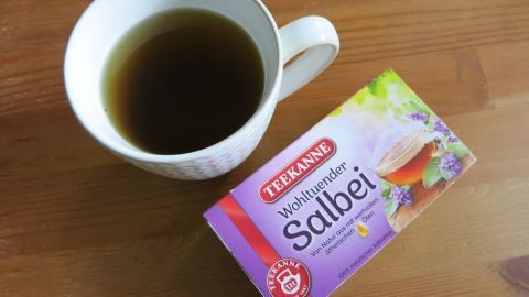 Eine Tasse mit Salbeitee steht neben einer Schachtel mit Salbeitee auf einem Holztisch, bereit für den Genuss gegen Fußgeruch.