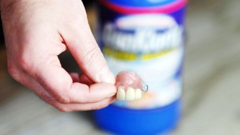 Eine Hand hält ein Gebiss, während im Hintergrund ein Reinigungsmittel steht, das für weiße Zähne genutzt wird.
