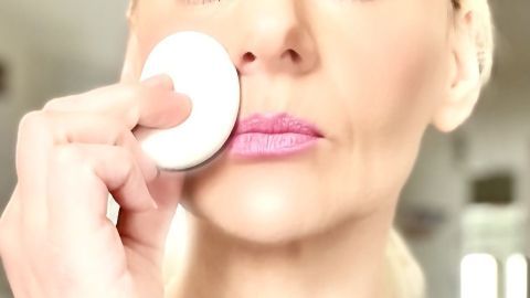 Eine Frau trägt Babypuder mit einem Schwämmchen sanft auf ihr Gesicht auf, während sie in einem hellen Raum steht.