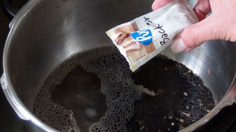 Ein Hand hält ein Päckchen über einen Topf mit verbranntem Inhalt, um ihn zu reinigen.