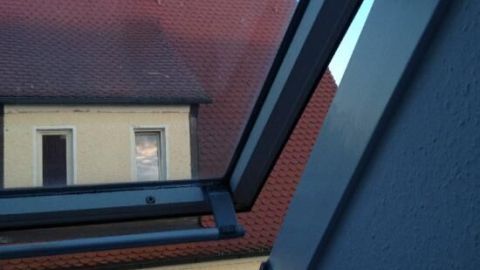 Ein offenes Dachfenster lässt frische Luft herein und bietet einen Blick auf ein rotes Dach und ein angrenzendes Gebäude.