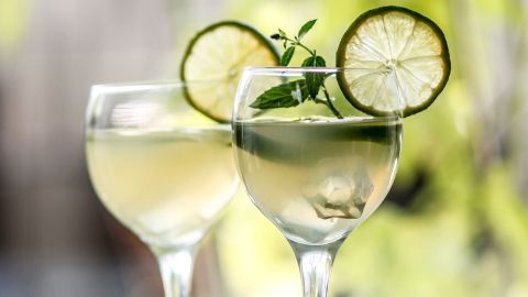 Zwei elegante Gläser mit einem erfrischenden alkoholfreien Hugo-Dink, garniert mit Limettenscheiben und Minze, stehen in einem sonnigen Garten.