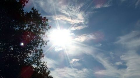 Eine strahlende Sonne steht hoch am Himmel, während Wolken sanft vorbeiziehen; sie deutet auf große Hitze hin.