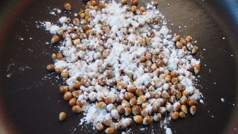 Popcornkörner liegen in einer Pfanne, bestreut mit Puderzucker, bereit zum Poppen. Süße Zubereitung in der Küche.