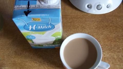 Ein Tetra Pak Milch steht neben einer Tasse mit braunem Kaffee auf einem Tisch, während ein Wasserkocher im Hintergrund sichtbar ist.