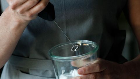 Eine Hand mit einem Schneebesen schlägt Sahne in einem transparenten Becher, um sie steif zu schlagen und vorzubereiten.