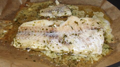 Ein Fischfilet liegt auf Backpapier, während es im eigenen Saft dünstet, umgeben von Kräutern und Zwiebeln.
