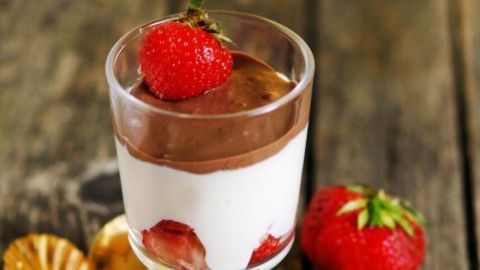 Ein Glas mit Erdbeeren in Creme und Schokoglasur steht auf einem rustikalen Holztisch, daneben liegen zwei goldene Löffel.
