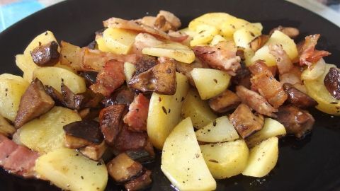 Gebratene Pilze und Kümmel-Speck-Kartoffeln liegen appetitlich auf einem schwarzen Teller, dampfend in einer Küche serviert.