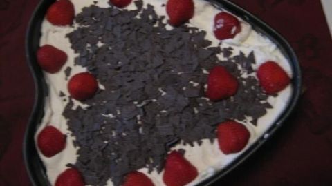 Eine herzförmige Erdbeer-Sahne-Schokotorte liegt auf rotem Stoff, dekoriert mit Schokolade und frischen Erdbeeren.