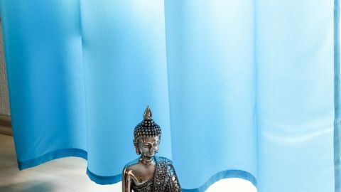 Ein blauer Vorhang hängt leicht und lässt Licht herein, während eine Buddha-Statue ruhig auf dem Boden sitzt.