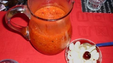 Krug mit roter Kirsch-Gazpacho steht auf rotem Tischtuch neben Schälchen mit Mandelscheiben und einer Kirsche in altem Esszimmer.