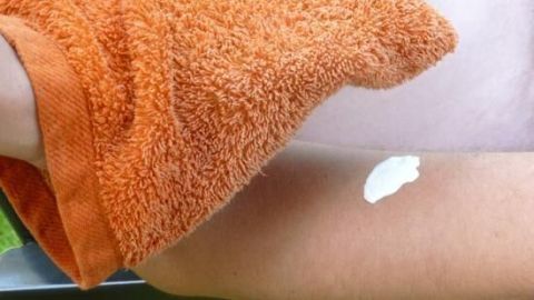 Ein feuchter Waschlappen trägt Sonnencreme auf einen Arm auf, während man auf einer Liege im Freien sitzt.