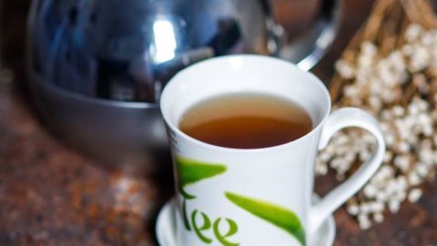 Eine Tasse grüner Tee steht vor einem glänzenden Kessel auf einer Holzoberfläche, umgeben von zarten Pflanzen.