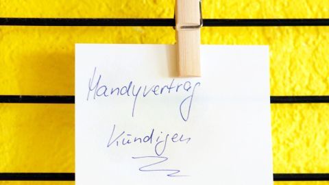 Ein Notizzettel mit "Handyvertrag" und "kündigen" ist an einer Schnur vor einer leuchtend gelben Wand befestigt.
