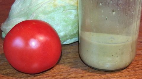 Ein roter Tomate und ein Glas mit Senf-Sauce stehen auf einem Tisch neben Kopfsalat, bereit für ein frisches Dressing.