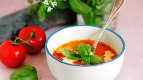 Eine schlichte Schüssel mit geeister Tomatensuppe wird von frischen Tomaten und Basilikum umgeben, in einer hellen Umgebung.