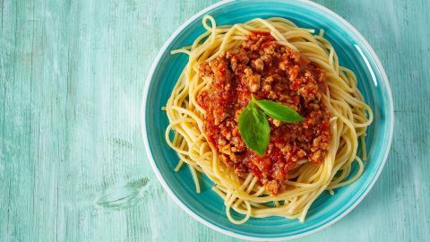 Spaghetti mit Hackfleischsoße auf einem bunten Teller, angerichtet auf grünem Holz. Ein frisches Basilikumblatt verziert das Gericht.