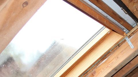 Ein Dachfenster ist geöffnet und zeigt verschmutztes Glas, umgeben von Holzbalken im Dachgeschoss.