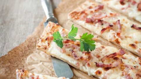 Ein Flammkuchen mit knusprigem Teig und Belag aus Schmand, Speck und Zwiebeln liegt auf einem Holzbrett, umgeben von Zwiebeln und Kräutern.