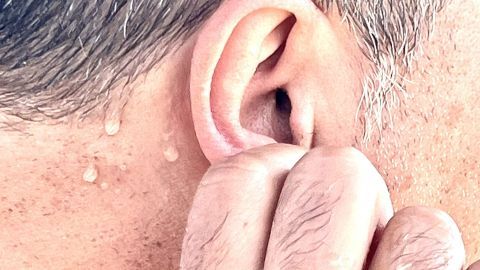 Die Ohrläppchen mit kaltem Wasser abreiben kann bei Hitze Abhilfe schaffen. Eine Person mit nassem Ohr greift sich mit der Hand ans Ohr, während sie sich in der heißen Umgebung abkühlt.