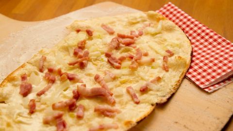 Ein frisch gebackener Flammkuchenboden liegt auf einem Holzbrett, umgeben von einem rot-weißen karierten Serviettenstück.