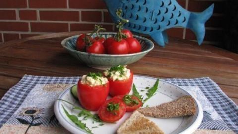 Rote Tomaten sind auf einem Teller mit Eiersalat gefüllt, umgeben von Scheiben Schwarzbrot auf einem karierten Tisch.