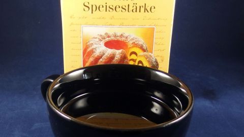Eine schwarze Schüssel mit feiner Speisestärke steht vor einer Packung, die das Produkt zeigt, auf einem blauen Untergrund.