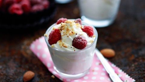 In einem klaren Glas befindet sich eine Himbeer-Krokant-Creme, die mit frischen Himbeeren und gehackten Nüssen garniert ist, auf einem rustikalen Tisch.