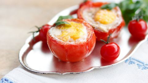 Zwei gefüllte Tomaten mit Ei liegen auf einer silbernen Platte, umgeben von kleinen Cocktailtomaten und frischen Kräutern.