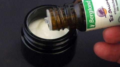 Eine Flasche Teebaumöl wird über eine Creme in einem eleganten, schwarzen Behälter gehalten, um sie zu mischen.