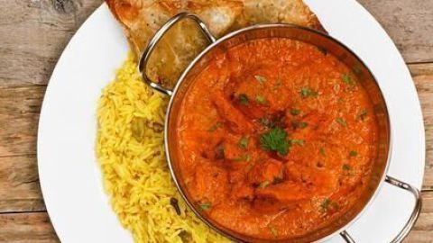 Ein roter Chicken Tikka Masala wird auf gelbem Reis serviert, umgeben von knusprigem Brot auf einem weißen Teller.