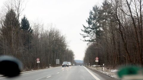 Eine Landstraße führt durch einen bewaldeten Bereich. Fahrzeuge fahren auf der Straße, während der Himmel bewölkt ist.