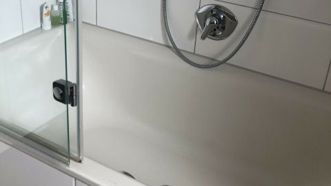 Eine moderne Badewanne mit Duschverkleidung ist sichtbar, die Wassertemperatur wird beschrieben. Die Umgebung wirkt klar und sauber.