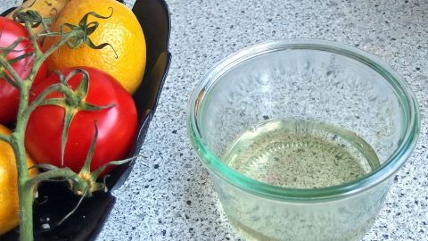 Ein Glas mit einer Flüssigkeit steht neben einem Obstkorb mit roten und gelben Tomaten auf einer modernen Küchenoberfläche.