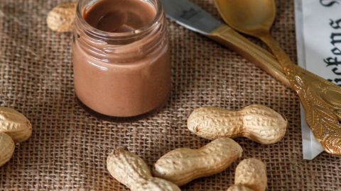 Ein Glas mit selbstgemachter Erdnussbutter steht auf einem juten Tuch, umgeben von ungeschälten Erdnüssen und Besteck.