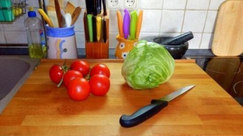 Auf einem Holzschneidebrett werden frische Tomaten und ein Kopf Salat mit einem scharfen Küchenmesser vorbereitet.