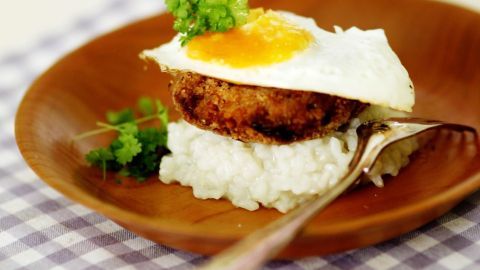 Ein Teller mit Loco Moco zeigt einen Burger auf Reis, garniert mit einem Spiegelei und frischen Kräutern, auf einem Holzteller.