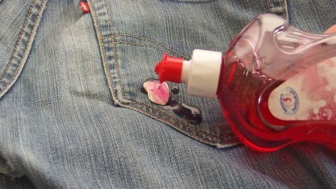 Ein Flasche Spülmittel wird auf einen Jeansstoff gegossen, um Kaugummi zu entfernen. Der Hintergrund zeigt einen unordentlichen Tisch.