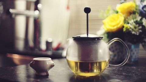 Eine Druck-Kaffeekanne mit goldgelbem Tee steht auf einer dunklen Arbeitsfläche, umgeben von frischen Blumen im Hintergrund.