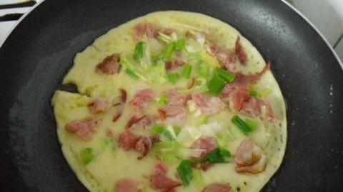 Ein Pfannkuchen brät in einer Pfanne, belegt mit Schinken und Frühlingszwiebeln in einer heimischen Küche.