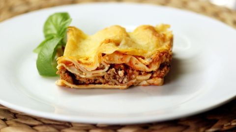 Ein Stück Lasagne liegt auf einem weißen Teller, während frisches Basilikum daneben dekorativ platziert ist.