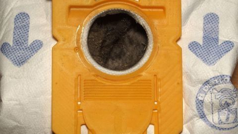 Ein gelber Staubsaugerbeutel liegt auf einem weißen Untergrund. Er zeigt eine Öffnung, aus der Staub sichtbar ist.